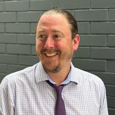 Dr. Mike Karlin from CSUDH, wearing a button-down shirt and tie, looking to the left in front of a gray brick wall