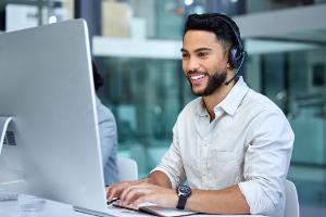 Happy young male sales agent talking to a customer via headset and working on computer station